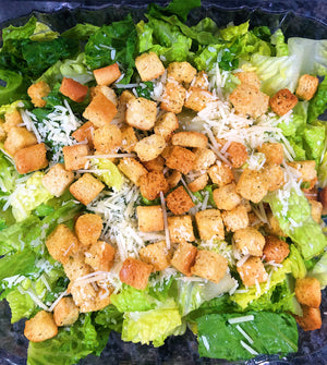 An image of a Caesar salad. The salad is served in a large bowl and consists of crisp romaine lettuce leaves. It is topped with shaved Parmesan cheese, croutons, and a creamy Caesar dressing. The salad is garnished with a lemon wedge and freshly ground black pepper. It presents a classic and appetizing combination of flavors and textures.